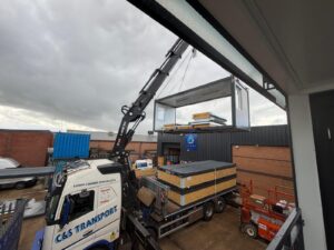 A lorry crane lifting a windowed modular unit above a flatbed carrying a second unit, in an industrial yard.