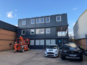 Two hi-vis workers in hard hats on an orange mobile lift beside a three-storey modular office building signed JSS&I Offices.