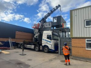 A lorry-mounted crane lifting a dark modular cabin above a stack of similar units, with three hi-vis workers coordinating.