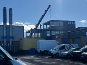 A crane lifting a grey windowed modular building above a roof, with a worker on the roof guiding it.