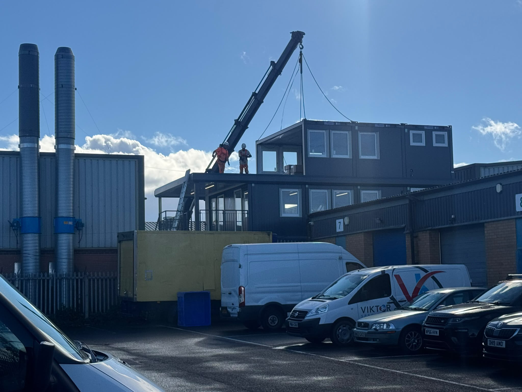 A lorry-mounted crane lifting a dark grey modular office unit into position on an existing stack of modular buildings at the JSS&I yard, with operatives in orange high-visibility clothing guiding the lift.