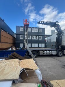 A worker in orange hi-vis on a raised red lift beside three stacked dark modular cabins, with a skip of timber offcuts below.